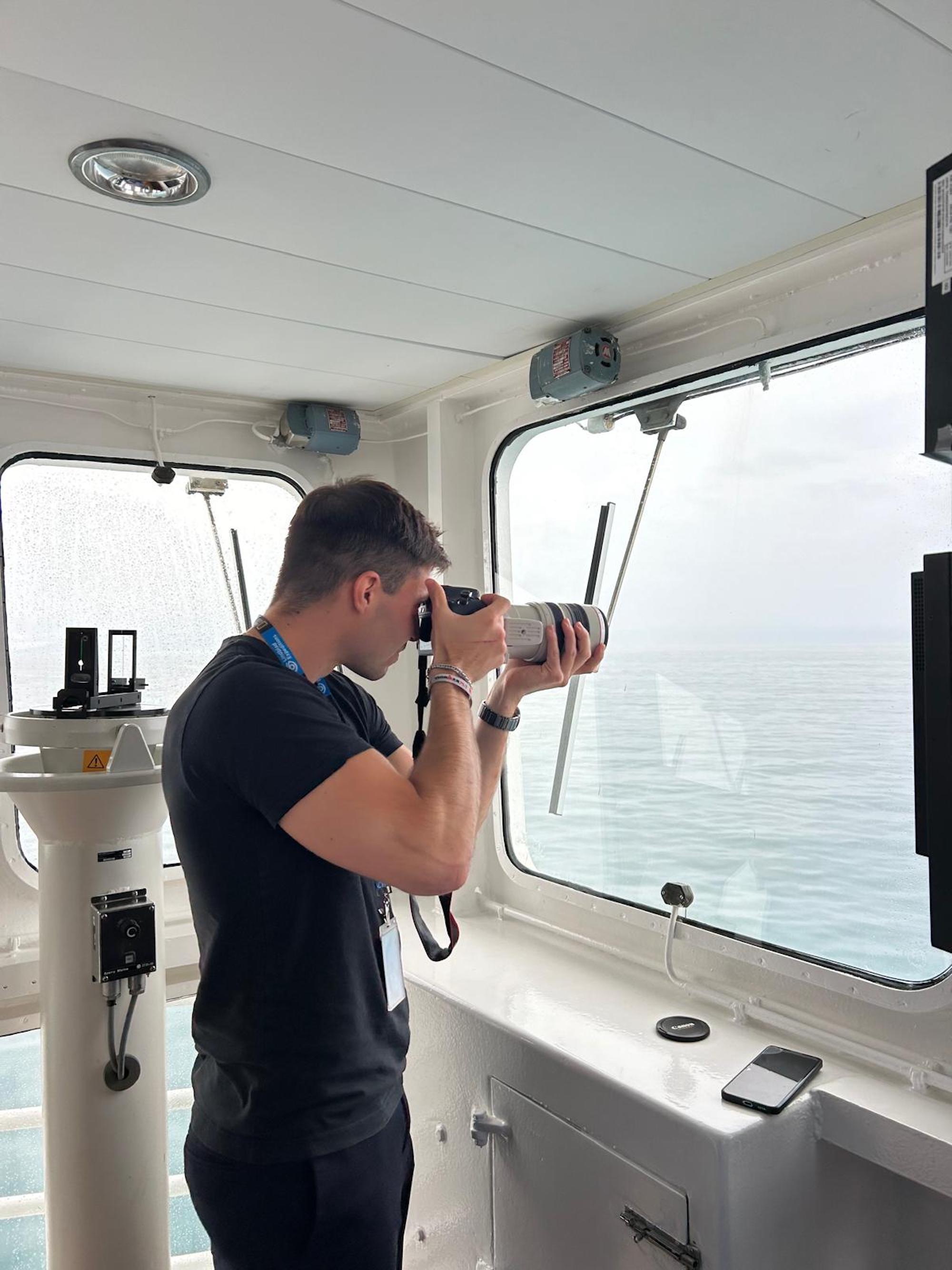 A researcher on a ship's bridge takes photos through a large camera, looking out at the ocean.