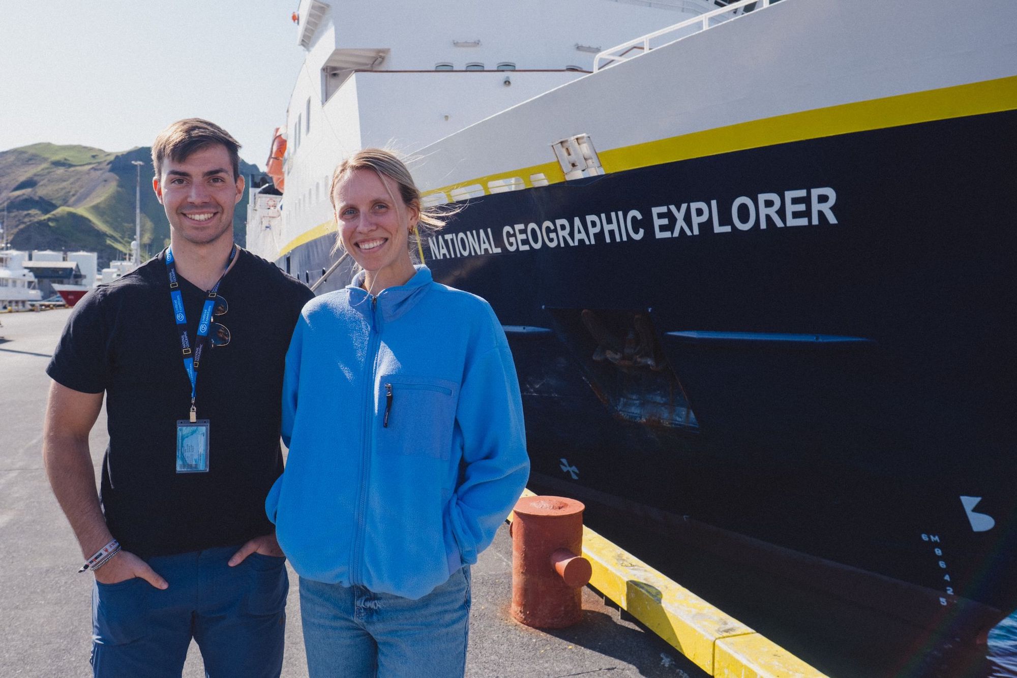 Two researchers pose together in front of the National Geographic Explorer ship at a harbor, smiling and wearing casual clothing.